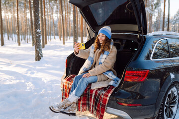 Selfie time. Young Woman sitting in the trunk of her car and taking selfie on a background of...