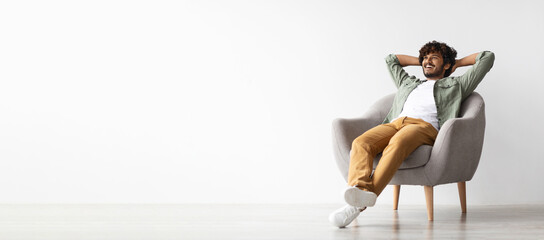 Relaxed curly bearded dark-skinned guy in casual outfit sitting at armchair with arms behind head,...