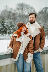 Love romantic couple lovestory. Brutal bearded man, bright red-haired girl woman in winter park. Romantic date, kissing, hugging. Walking, having fun. Stylish clothes, red sheepskin coat, jacket, hat