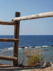 Obraz premium A simple wooden fence stands near a rocky coastline, offering a partial view of the calm blue ocean under a clear sky. Dry brush grows in the foreground.