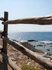 Fototapeta premium A rustic wooden fence runs along a rocky shoreline. The calm blue sea shimmers under a bright sun. Sunlight casts shadows on the weathered wood. The scene suggests a peaceful coastal location.