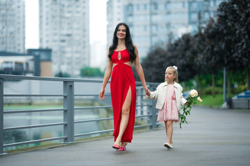 Obraz premium Woman in a Red Dress Walks Hand in Hand With Young Girl in a City Park