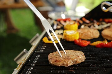 A metal spatula is being used to flip a burger patty on a grill. The burger is cooking alongside other grilled items, including corn on the cob, bell peppers, and mushrooms.