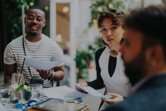 Three diverse professionals engaged in a collaborative meeting, discussing projects and strategies with documents in an urban coffee shop environment.