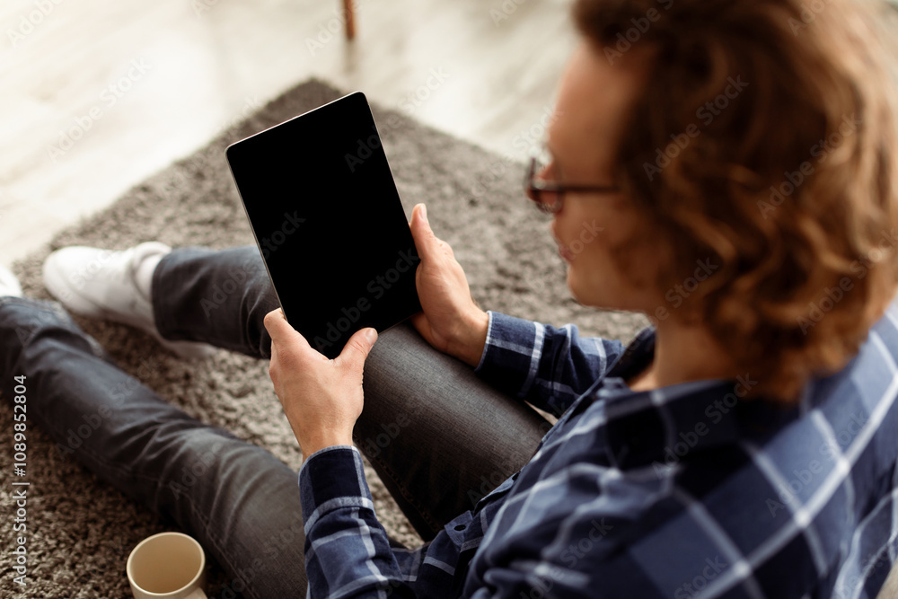 Poster Hipster Guy Using Digital Tablet Computer With Empty Screen Browsing Internet Sitting On Floor At Home. Website Advertisement Concept. Selective Focus, Rear View, Mockup
