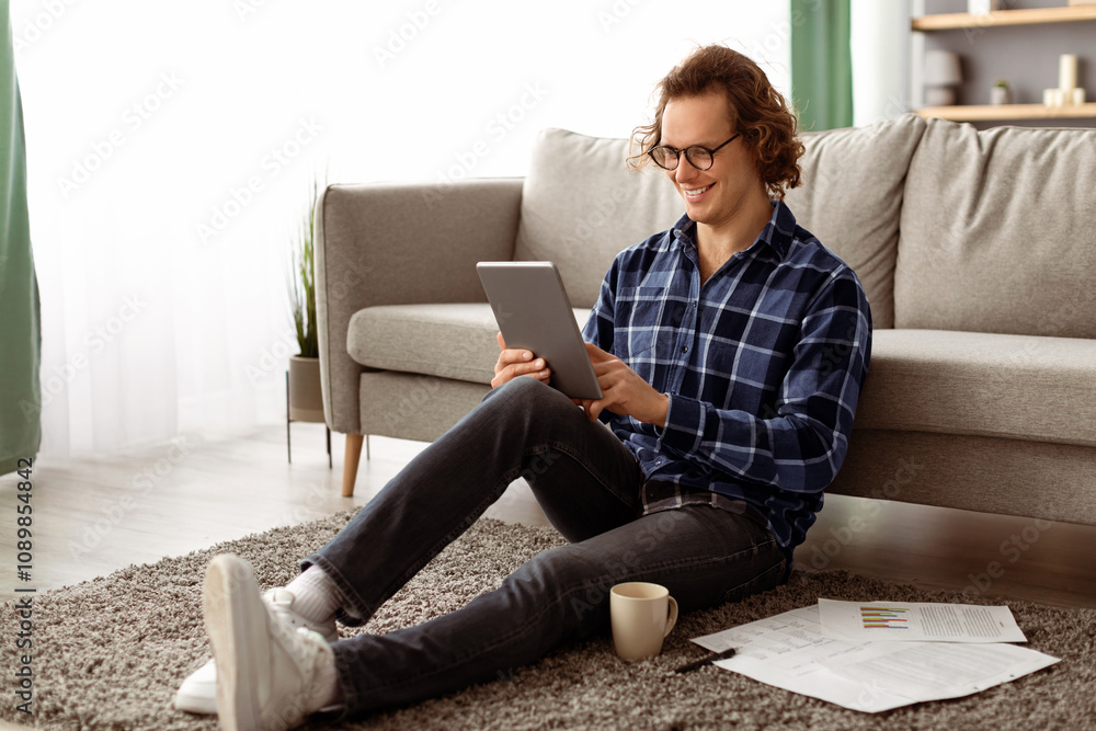 Poster Cheerful Man Using Tablet Computer Browsing Internet Sitting On Floor At Home. Guy Watching Movie Indoor. Application Or Website Advertisement, Gadget And Technology Concept. Side View