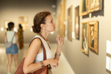 Side view of young intelligent woman looking at exhibition of paintings in modern museum