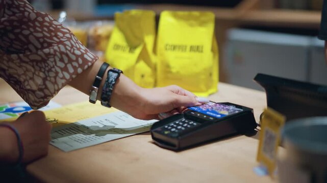 Close-up of person in vintage outfit holding card over a card reader to make payment, with wrist adorned with bracelets and a watch, yellow coffee bags and menu papers are visible on table