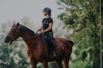 A woman dressed in equestrian gear rides a brown horse in a serene outdoor setting, surrounded by greenery. She appears focused and calm, enjoying the connection with the animal.