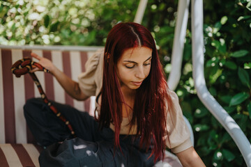 A girl with striking red hair is seated on a striped swing, holding a snake. She appears calm and...