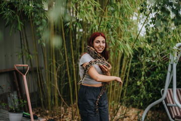 A smiling girl with red hair holding a large snake in a lush garden setting. The scene conveys a...