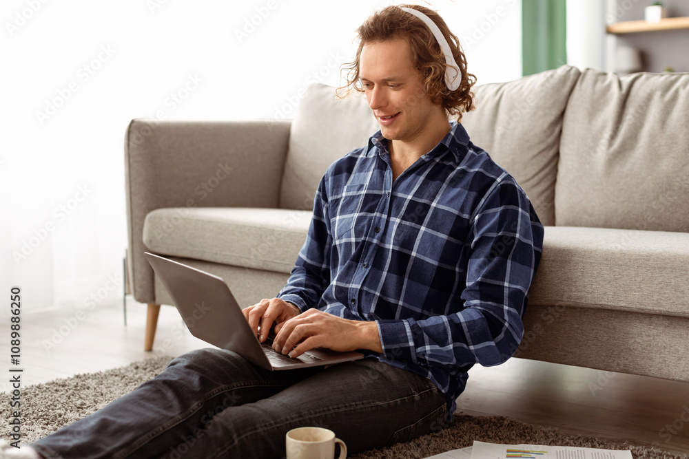 Poster smiling man in headphones using laptop making video call or working online sitting on floor at home.