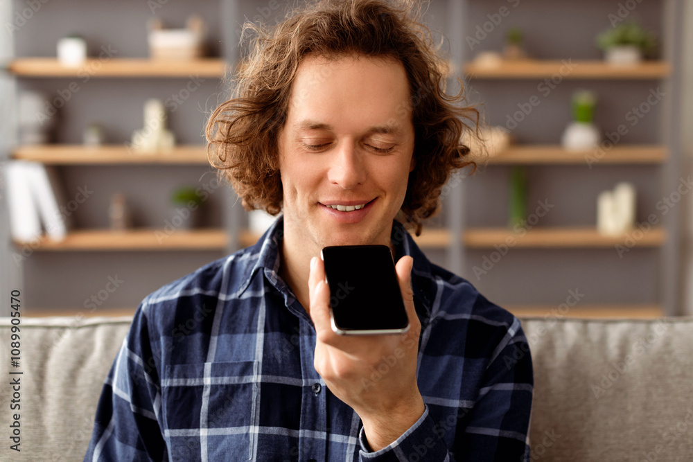 Poster guy using voice assistant on mobile phone sitting on sofa at home. long-haired millennial man talkin