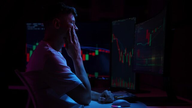 Stressed crypto trader touching head with hands while looking at multiple computer monitors showing falling stock market charts in dark room illuminated by blue neon lights. Shooting in slow motion.