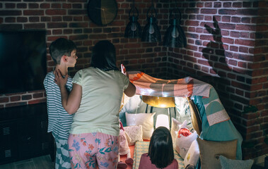 Back view of mother and two children having fun playing with shadow puppets over brick wall at home...