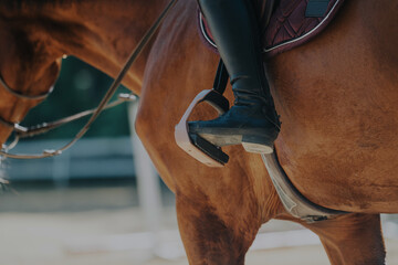 A detailed shot showcasing a rider's foot in stirrups, set against a brown horse. This image...