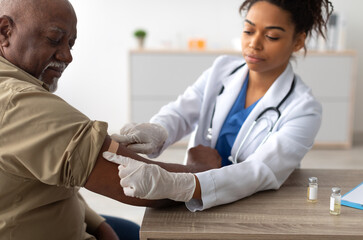 Close up of black female doctor in protective gloves applying sticking patch to patient's arm after...