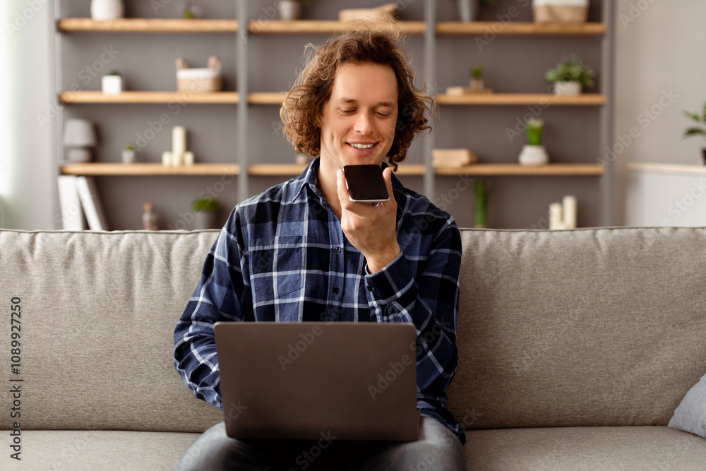 Aufkleber Man Using Voice Assistant On Smartphone Working On Laptop Sitting On Couch At Home. Modern Hipster Guy Talking With Loudspeaker Mode Holding Phone Near Mouth. Voice Search Mobile Application