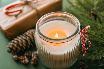 A lit candle in a festive glass holder surrounded by pinecones, evergreen branches, a wrapped gift,...