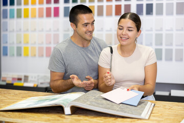 Obraz premium Smiling young woman and man looking for paints for house renovation in local construction hypermarket, talking friendly while browsing large catalog with coloring samples ..