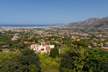 Naklejka premium Panoramic view of sprawling cityscape of Palermo in Italy framed by mountains and lush greenery, with clear blue sky and distant ocean in the background