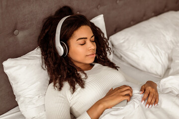 Rest And Relax Concept. Closeup portrait of calm black woman lying in bed, listening to music,...