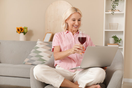 Mature woman with wine and laptop having online date in armchair at home