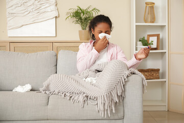 Ill African-American girl with thermometer and tissues sneezing on sofa at home