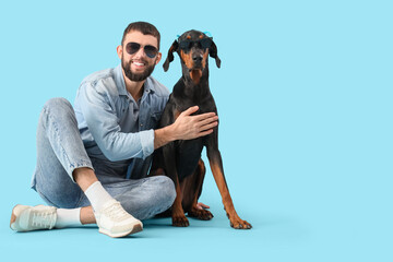 Young man with Dobermann dog in sunglasses sitting on blue background