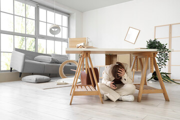 Young woman hiding under desk in living room during earthquake