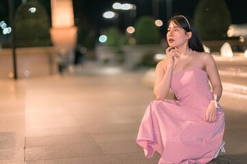 Naklejka premium Elegant young woman in a pink evening gown poses gracefully at night near a fountain, surrounded by soft lights and a serene atmosphere, exuding timeless beauty and charm.