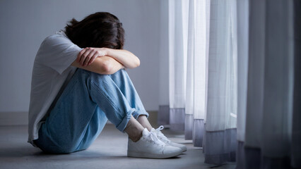 A person sits on the floor with their knees drawn up to their chest, their head lowered, and their arms loosely wrapped around their legs