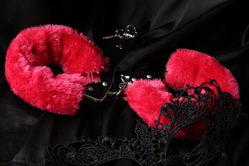 Red fluffy handcuffs, keys and lace mask on black fabric, closeup