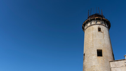 A fragment of an old lighthouse. A tall dilapidated cylindrical tower against a clear blue sky. Open dark window openings. Mauritius. Île aux Fouquets