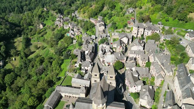 Drone aerial view in France countryside small old medieval town and a cathedral surrounded by a green mountain forest flying over in Conques