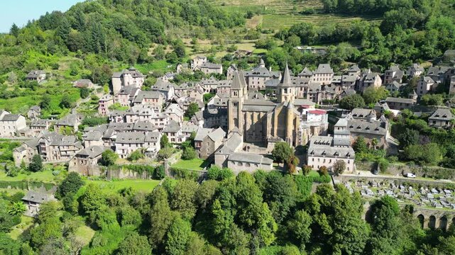 Drone aerial view in France countryside small old medieval town and a cathedral surrounded by a green mountain forest circling around in Conques