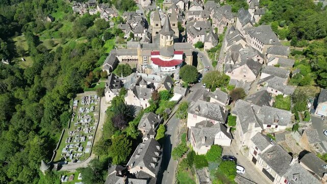 Drone aerial view in France countryside small old medieval town and a cathedral surrounded by a green mountain forest flying over in Conques