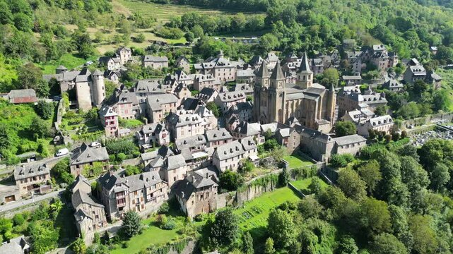 Drone aerial view in France countryside small old medieval town and a cathedral surrounded by a green mountain forest circling around in Conques