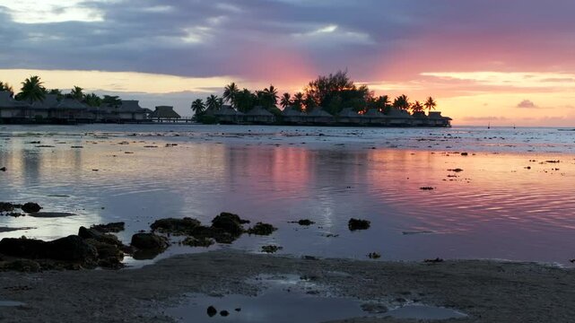 Stunning vibrant pink orange sunset Bungalow villas islands Cooks Opunohu Bay Moorea French Polynesia aerial drone clouds lagoon coral reef Pacific Ocean low tide beach slowly slide left motion