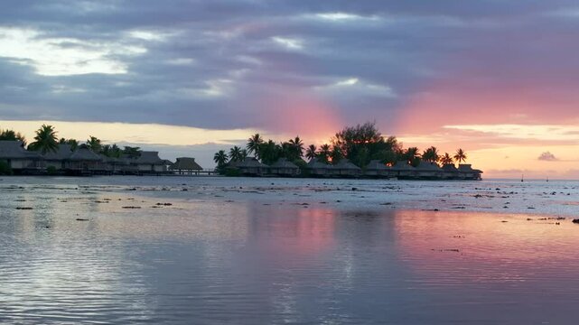 Stunning vibrant pink orange sunset Bungalow villas islands Cooks Opunohu Bay Moorea French Polynesia aerial drone clouds lagoon coral reef Pacific Ocean low tide beach slow slide right pan up motion