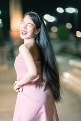 Obraz premium Young woman laughs joyfully while wearing a light pink dress, posing confidently against a blurred city backdrop at night, capturing the essence of happiness and glamour.