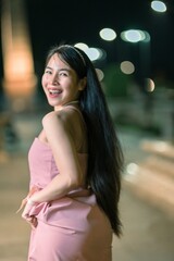 Fototapeta premium Joyful woman in a pink dress poses confidently at a vibrant night setting, showcasing her long hair and radiant smile against a backdrop of glowing lights.