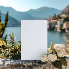 Blank Wedding Invitation Mockup: A pristine white card mockup rests on a stone ledge, adorned with white roses and greenery, against a breathtaking backdrop of an Italian lake and mountain scenery.  