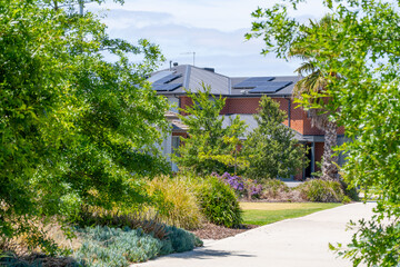 A footpath lined with lush green plants and trees in a public park leads to residential suburban...