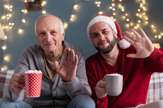 Senior man and his adult son in santa claus hat waving hello to camera sitting on sofa drinking cocoa with marshmallows on christmas morning. Merry christmas and happy new year
