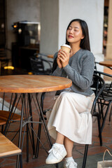 A beautiful Asian woman in cozy clothes enjoys coffee at a shop, smelling it with her eyes closed.