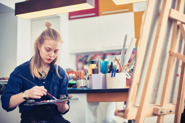 Young woman painting on canvas in a bright studio filled with art supplies at midday