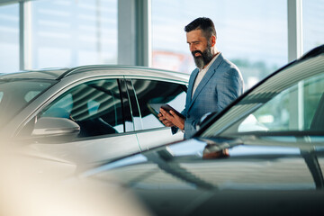 Man in suit examining car in dealership with digital tablet in hand.