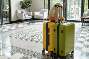 Bright green suitcase with a hat on handle standing in spacious, elegantly designed hotel lobby with patterned tile floor and stylish furniture all around