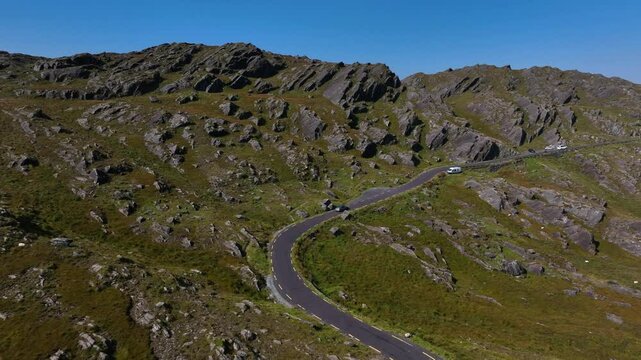 Healy Pass, County Cork, Ireland, September 2024. Drone ascends while following a car driving along single lane road towards the summit of the mountain bordering County Kerry under a clear blue sky.